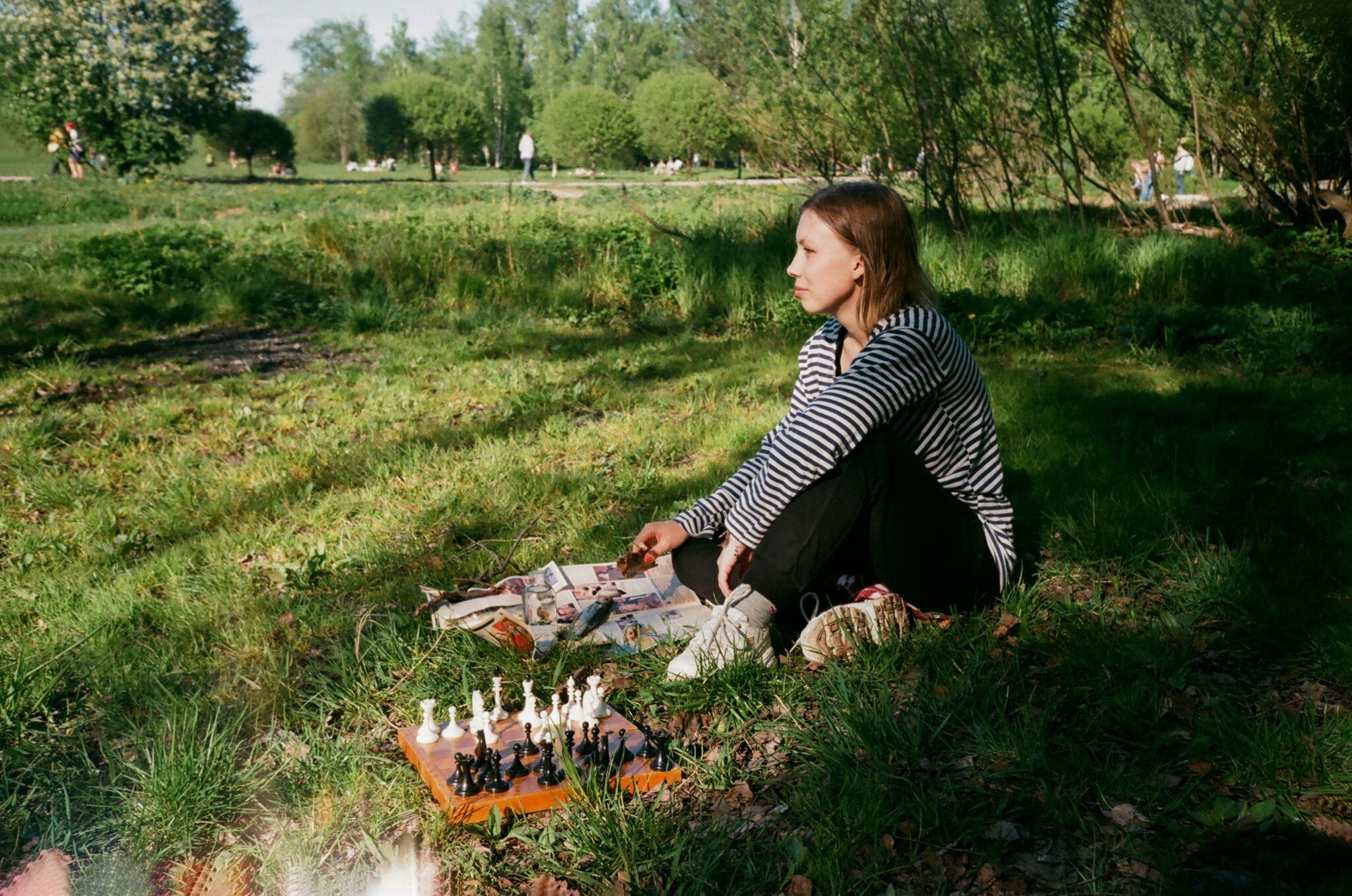 Full body side view of thoughtful young slender female in casual clothing looking ahead while playing chess in park immersed in greenery in sunlight